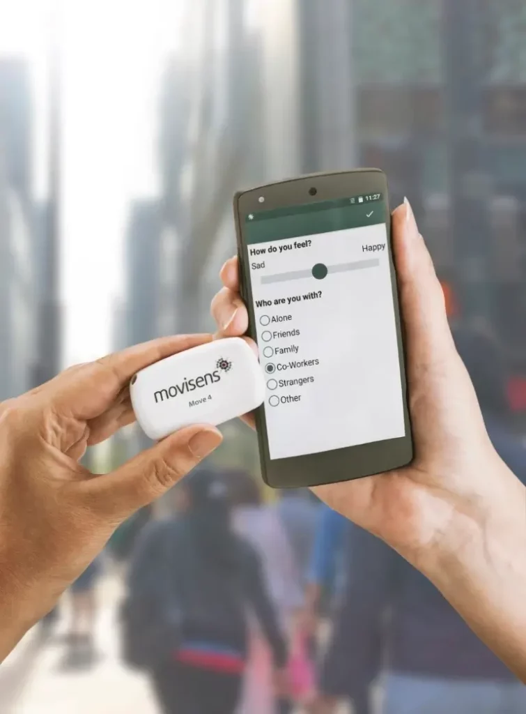 Person holding a movisens Move 4 wearable sensor in one hand and a smartphone displaying an ecological momentary assessment (EMA) survey screen in the other. The survey asks about current mood and social context, with options for “Sad” to “Happy” and “Alone,” “Friends,” “Family,” “Co-Workers,” “Strangers,” and “Other”
