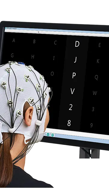 Person wearing a white EEG cap with electrodes connected to a computer monitor displaying a brain-computer interface (BCI) speller grid with letters and numbers highlighted for selection