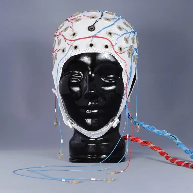 Black mannequin head wearing a white EEG cap with multiple electrodes and color-coded wiring, designed for MEG (magnetoencephalography) or EEG recordings in a research setting