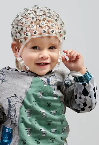 Young child wearing a BrainVision R-Net EEG cap with sponge-based passive electrodes, smiling and looking at the camera