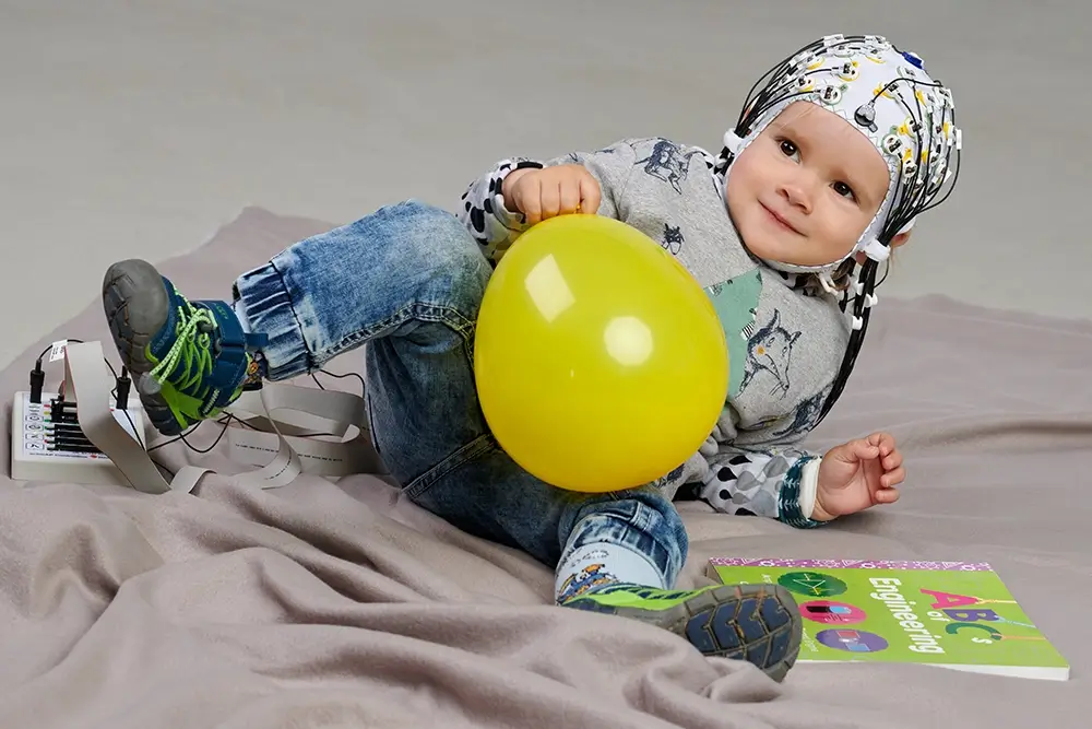 Toddler wearing an EEG cap with actiCAP snap electrodes, holding a yellow balloon while sitting on a soft surface, connected to an amplifier device, with an ABC engineering book nearby