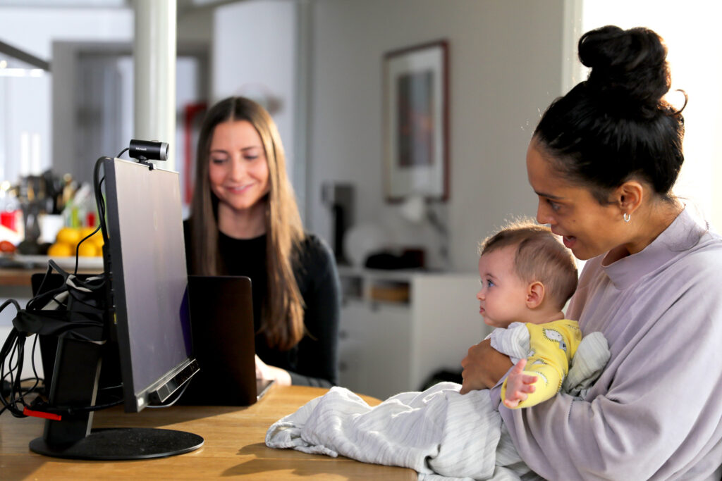 A caregiver holds a baby in their lap while they both fact a computer screen that is fitted with an eyetracker (Tobii Pro Fusion) and scene camera. A researcher is visible in the background using a different computer that will control the images shown on screen to the baby and caregiver.