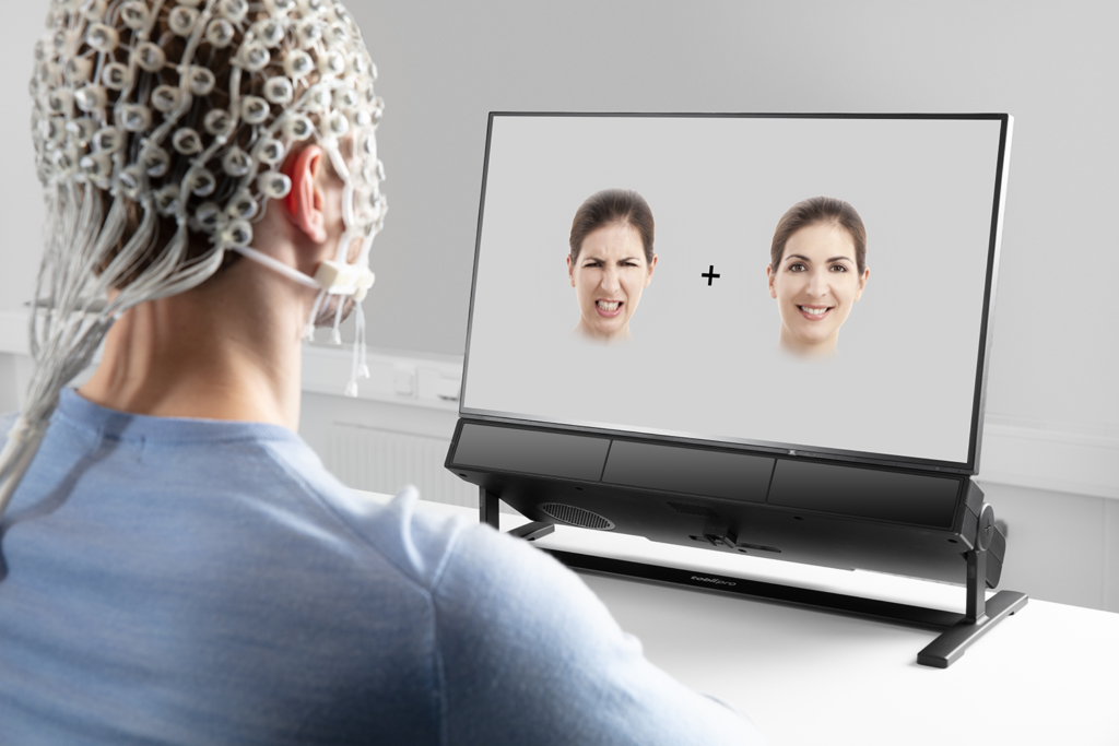 We are looking over the shoulder of a person in a blue shirt while they wear an R-Net (saline-sponge EEG net) and sit in front of the Tobii Pro Spectrum stationary eyetracking unit. On screen two faces with different emotions are visible and the eye tracker is monitoring where the person's gaze attendes on screen.