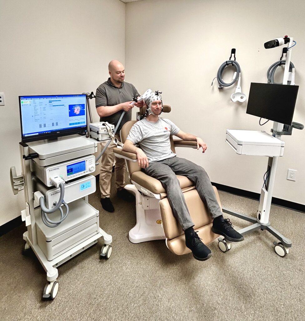 A male participant wearing an active electrode EEG cap is seated in a large brown and white treatment chair. Behind him stands a researcher holding a TMS coil. To their left is a neuronavigation setup and to their right is the TMS system and its controlling computer.