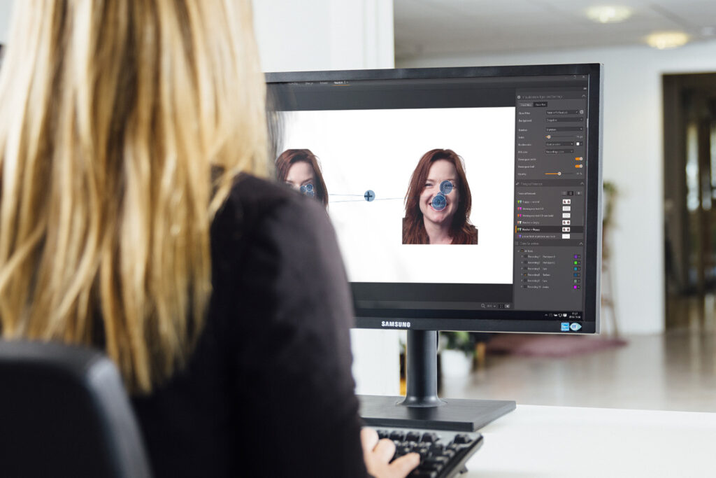 A researcher with long blond hair sits in front of a computer that is running Tobii Pro Lab software. On the software, they are reviewing the eye tracking data from someone doing a task that showed different faces. The software interface buttons and gaze position can be seen on screen.